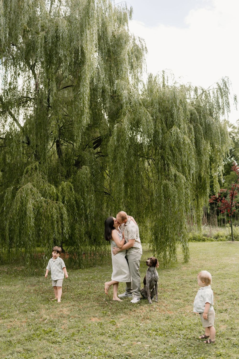 Family kissing under willow tree with two children and a dog on green lawn.