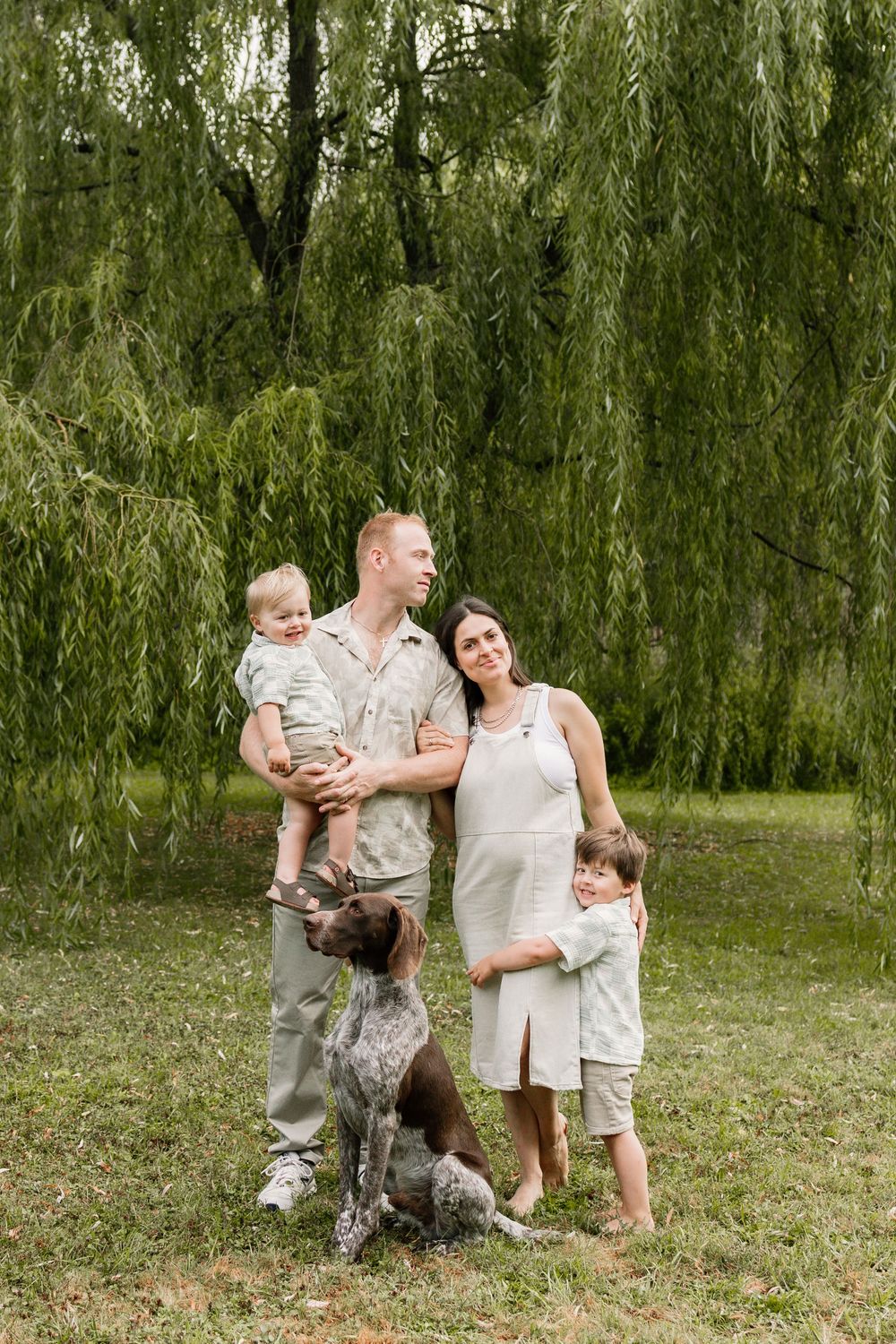 Family with two kids and a dog posing outdoors under a weeping willow tree on a sunny day.