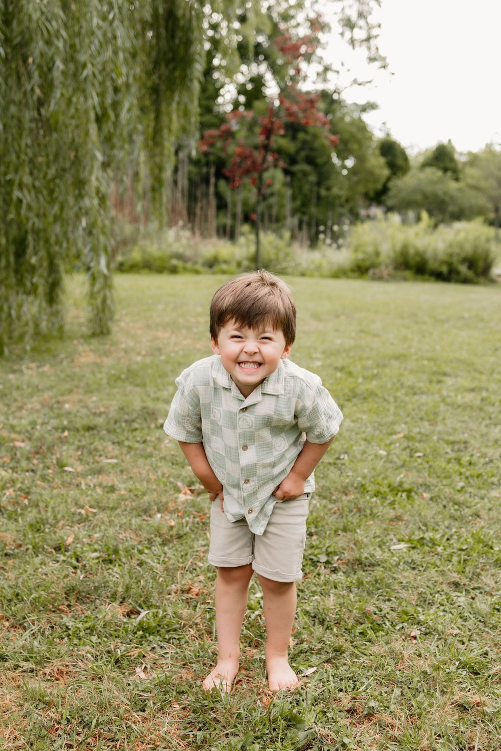 A smiling young boy in a green plaid shirt and khaki shorts stands barefoot on grass near a willow tree.