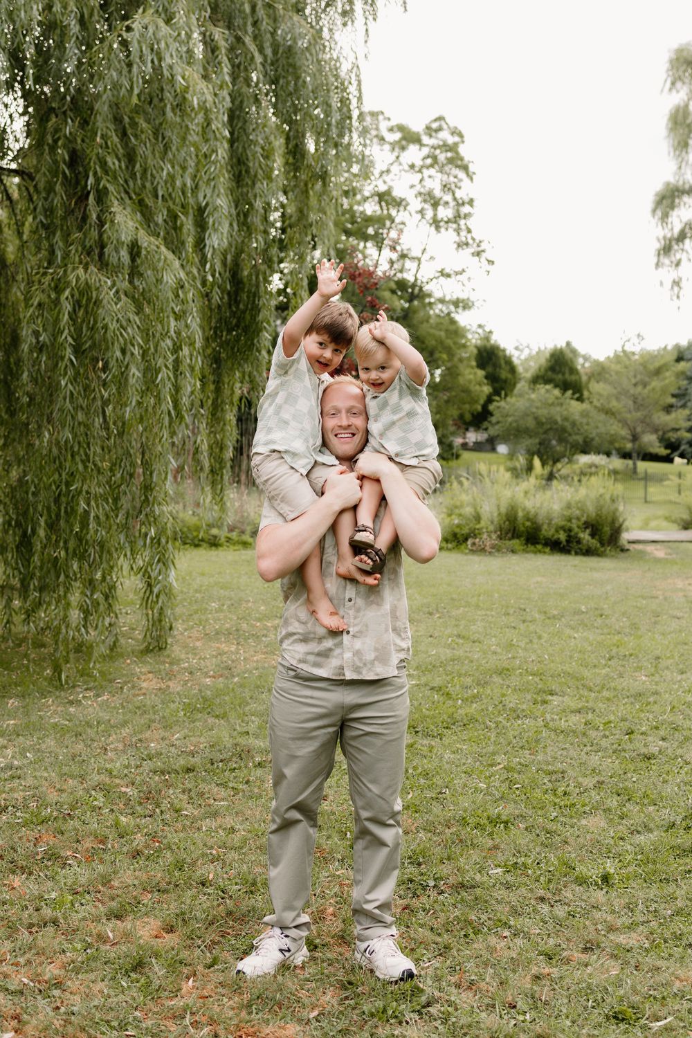 A smiling man carries a young child on his shoulders in a lush green park near a willow tree.
