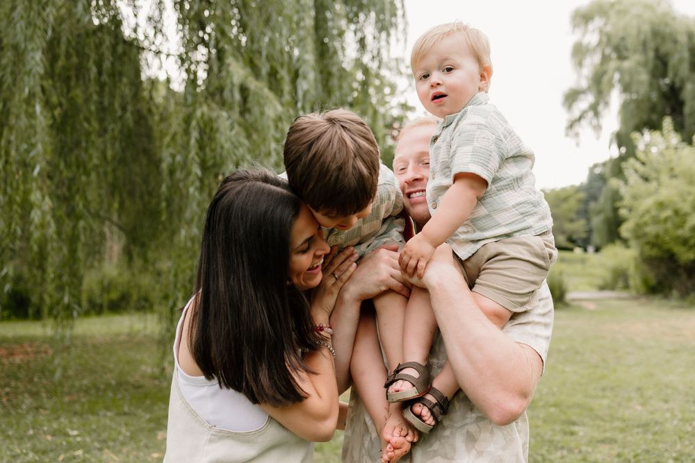 Happy family with two parents and toddler boy laughing together outdoors near a weeping willow tree.
