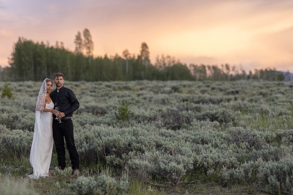 A bride and groom embrace in a sagebrush field at sunset with a glowing pink sky and tree line behind them.