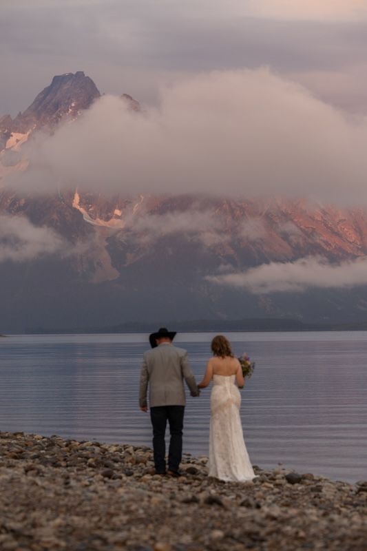 Bride and groom holding hands at lakeside with misty mountain backdrop at sunset during outdoor wedding ceremony.