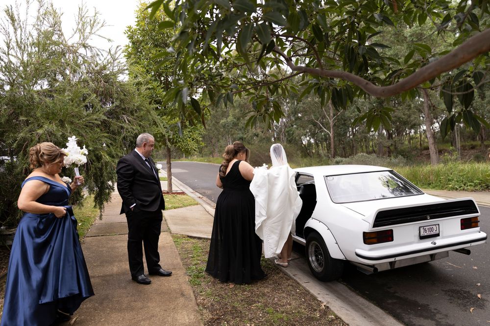 Bride in white gown entering a classic white car with wedding party in blue and black attire on a tree-lined driveway.