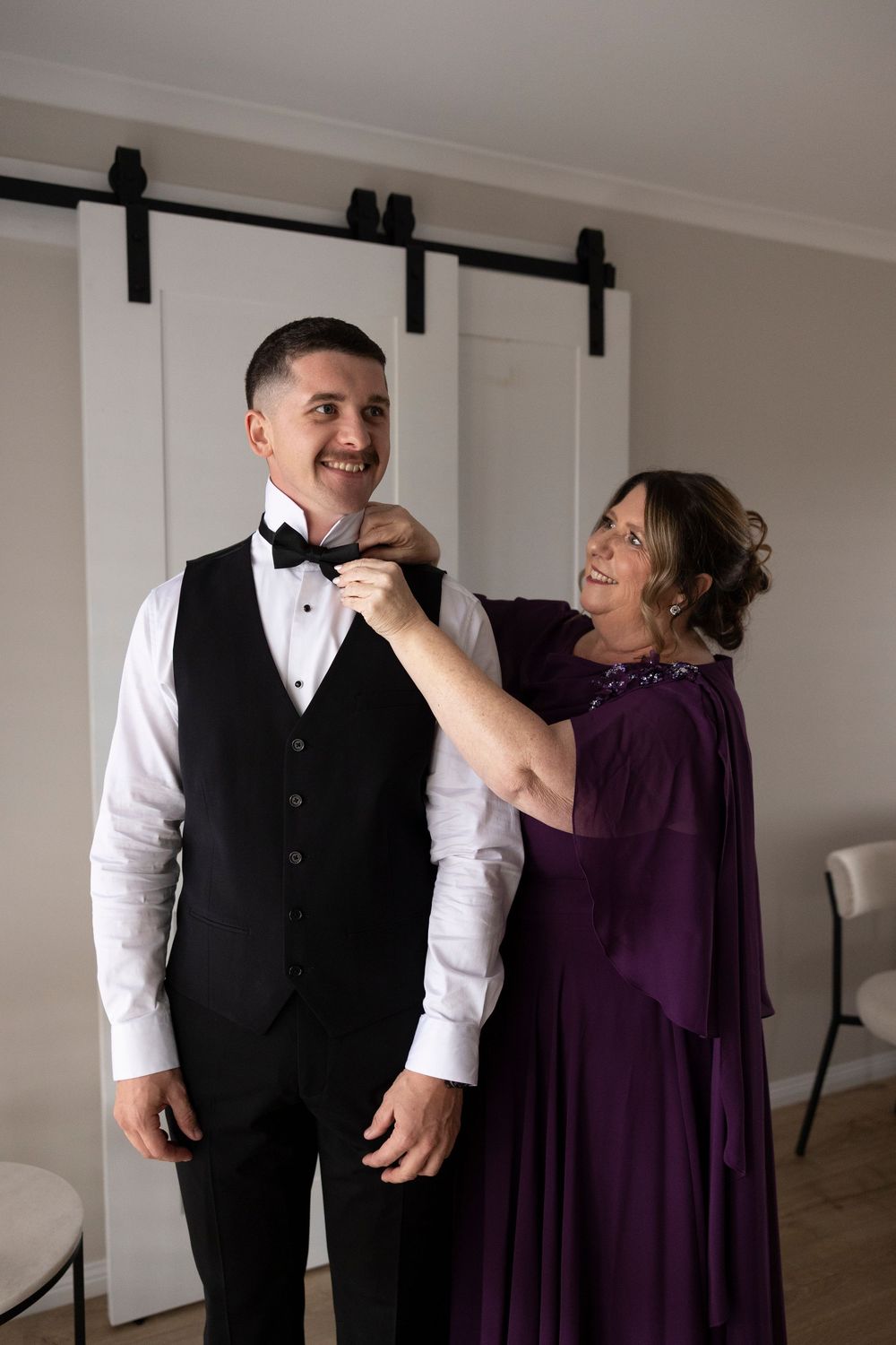 Smiling groom in black tuxedo vest gets bow tie adjusted by woman in purple gown indoors.