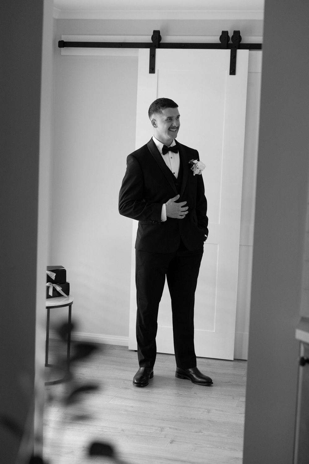Black and white photo of a groom in a tuxedo with boutonniere smiling in a bright room with barn doors.
