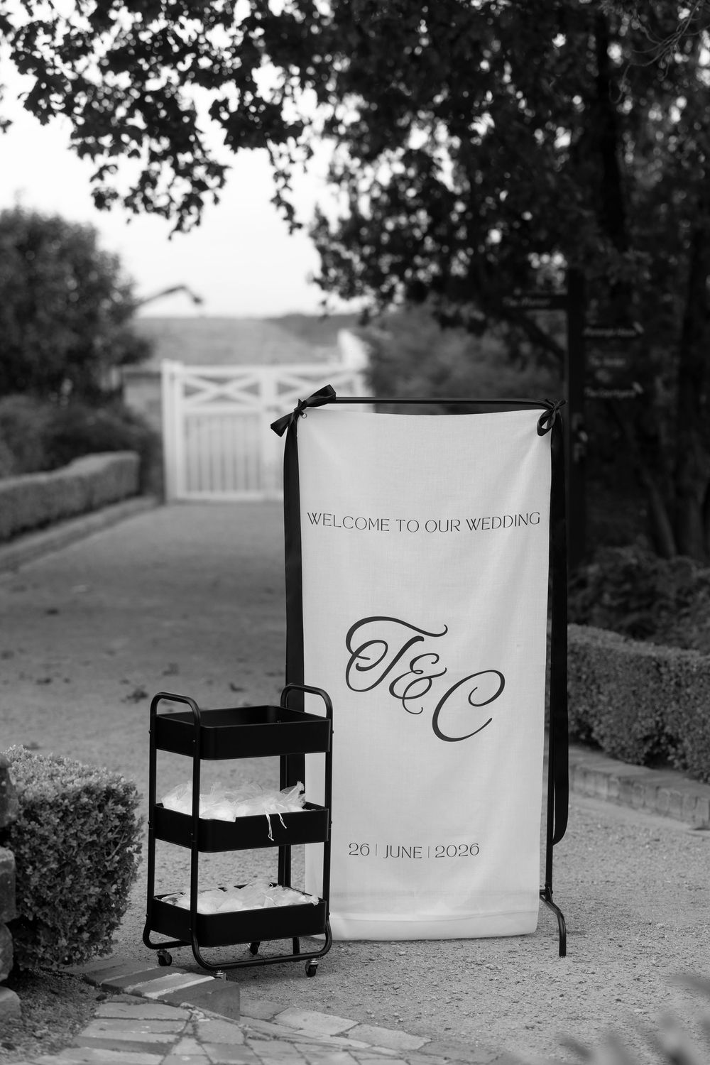 Black and white wedding welcome banner with T&C monogram dated 20 June 2020 displayed outdoors.