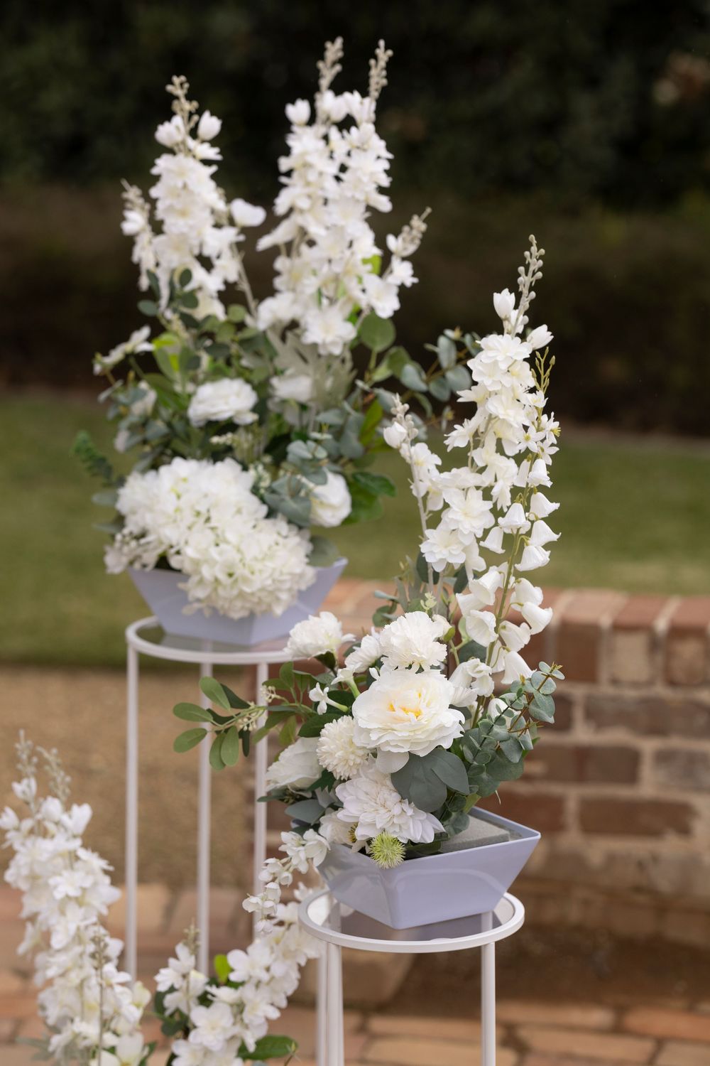 White floral arrangements with hydrangeas and delphiniums on white stands at an outdoor wedding ceremony.
