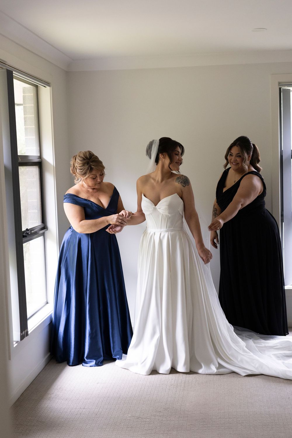 Bride in white strapless gown flanked by two bridesmaids in navy and black dresses during wedding preparations.