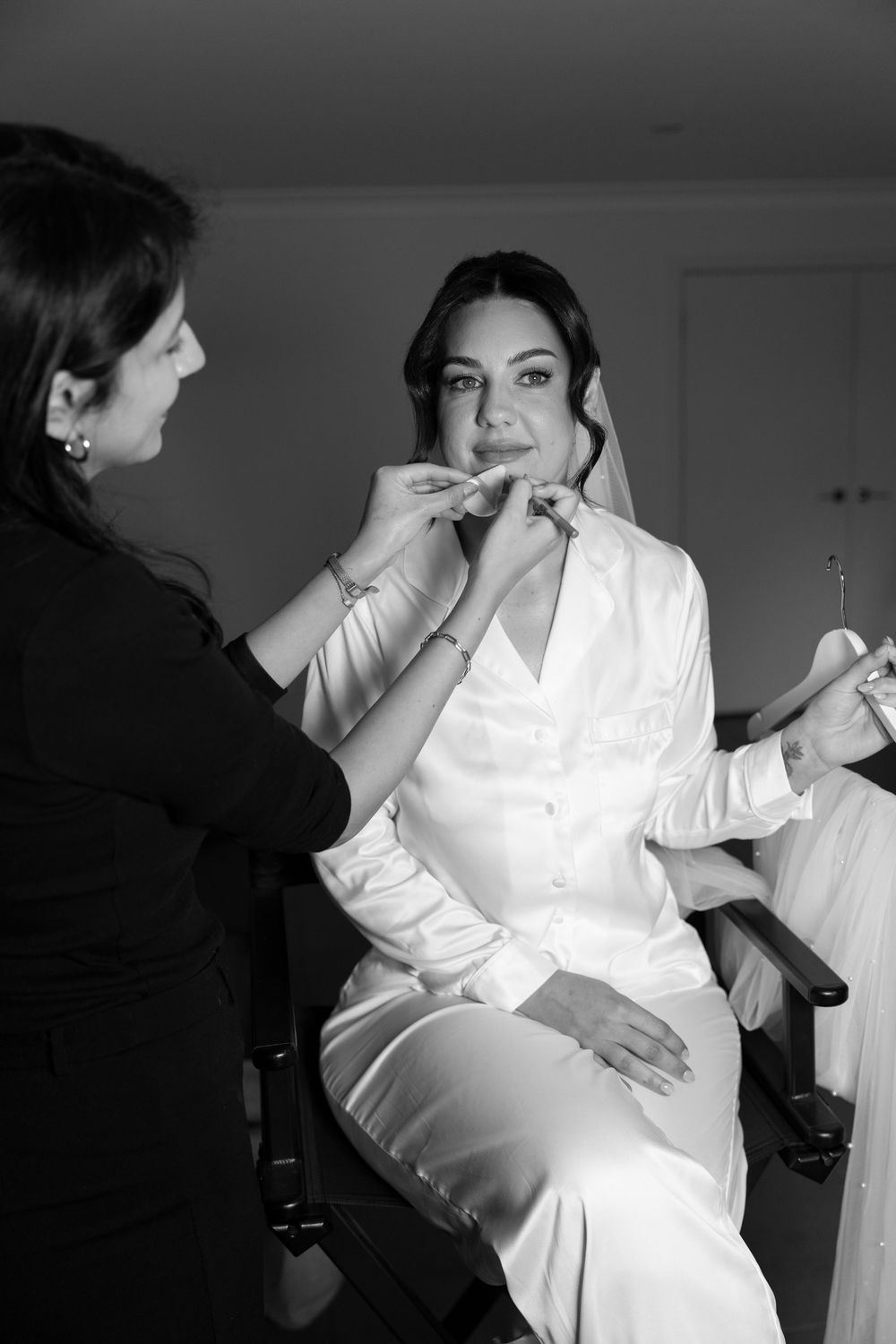 Black and white photo of a bride in silk robe getting makeup applied by a makeup artist before her wedding.