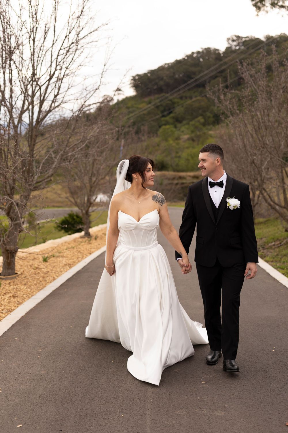 Bride and groom walk hand-in-hand on a tree-lined path, smiling at each other in wedding attire.