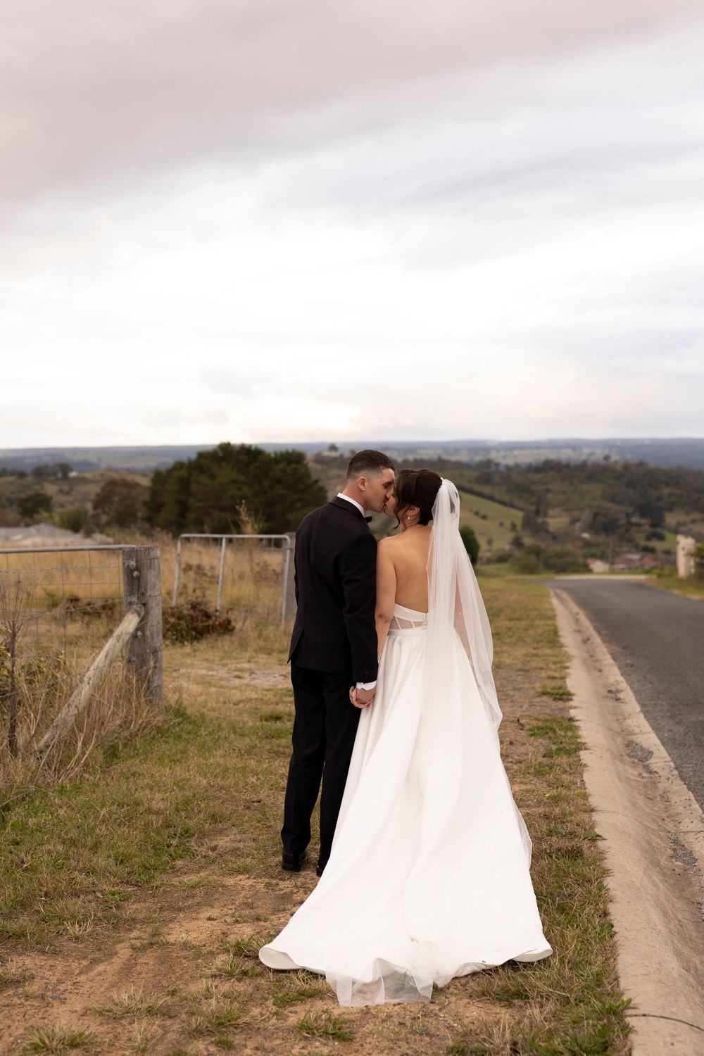 Bride and groom share a romantic kiss on a rural road, bride wearing a flowing white gown with veil.