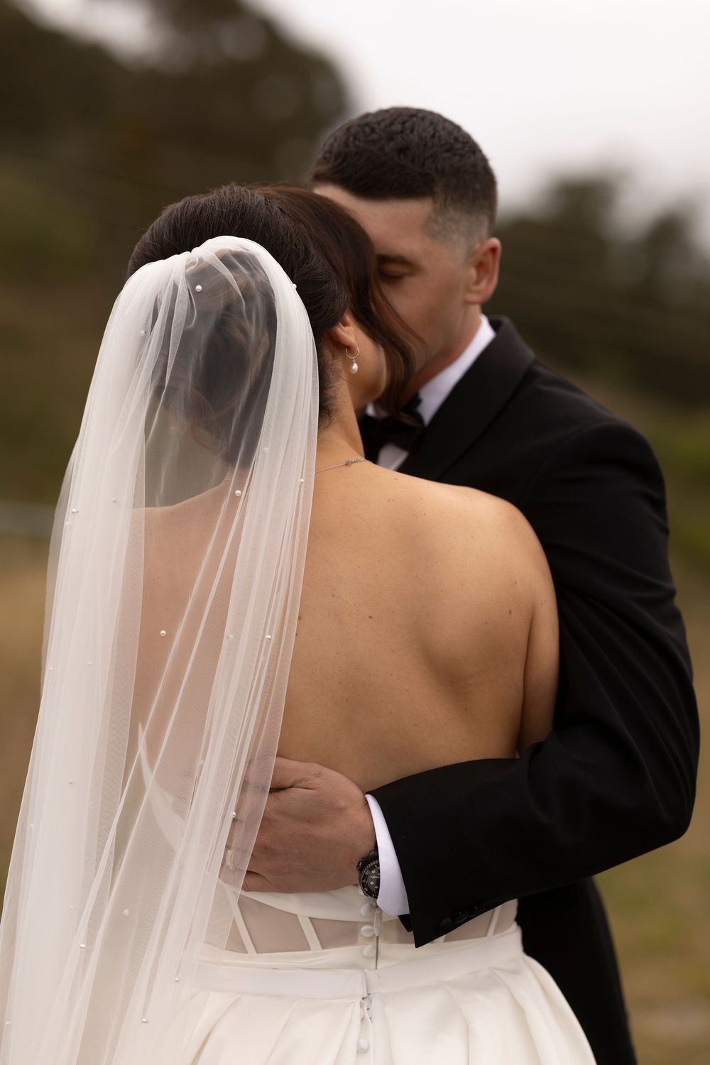 A bride and groom share a romantic embrace outdoors, with the bride wearing a white gown and veil.