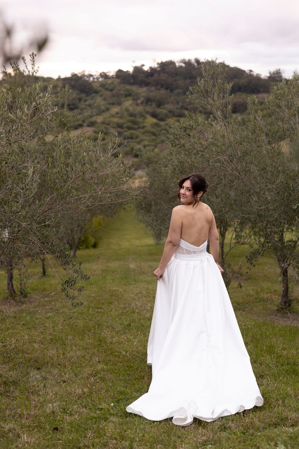 Bride in backless white gown stands among olive trees in lush green orchard, glancing over shoulder.