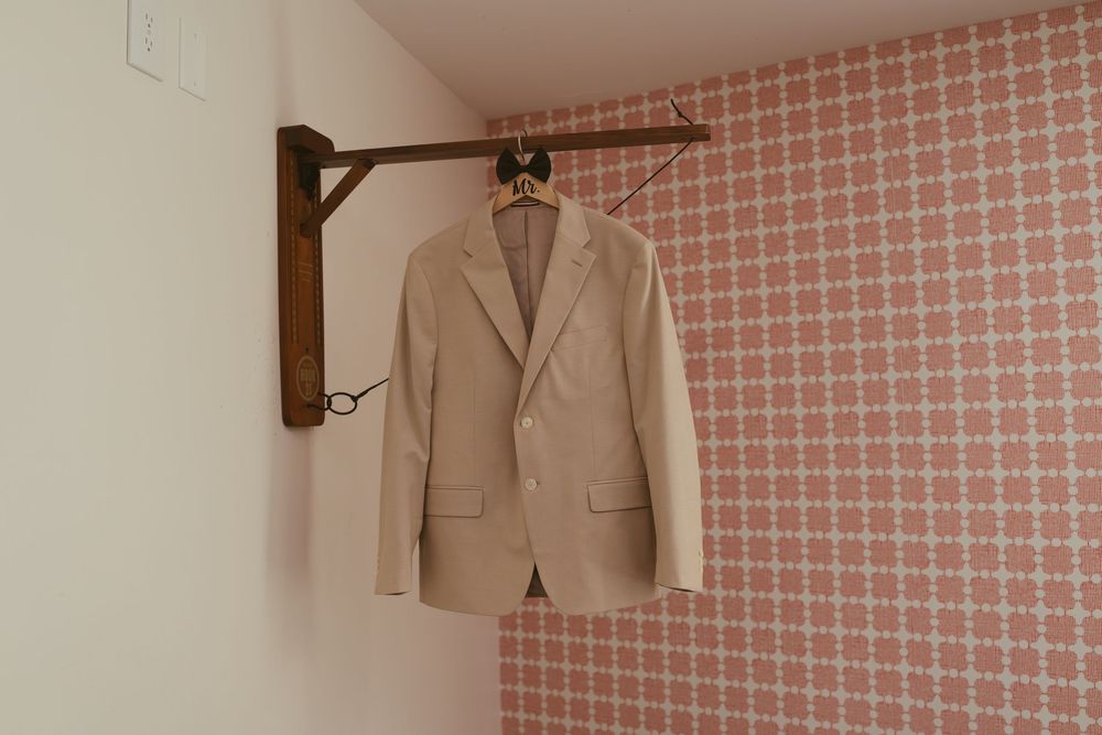 A beige blazer hanging on a wooden wall-mounted rack against pink geometric patterned wallpaper.