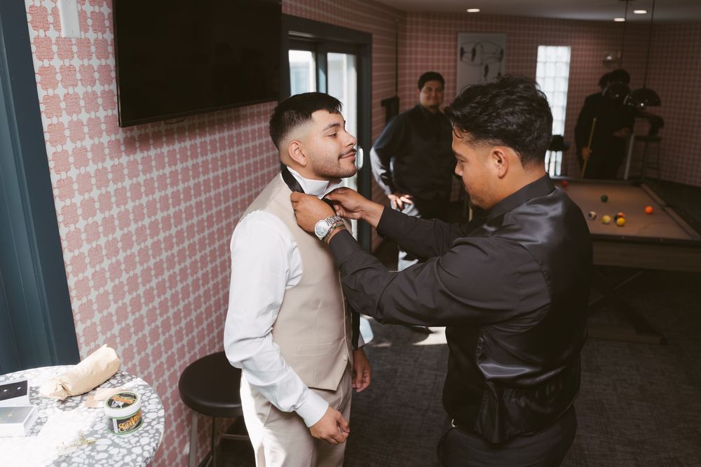A man in a white shirt and vest gets his tie adjusted by another man in a black jacket indoors.