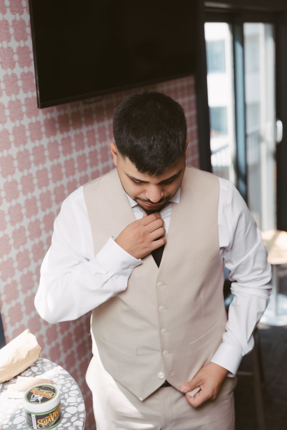 A groom in a white shirt and beige vest adjusts his tie while getting ready for his wedding.