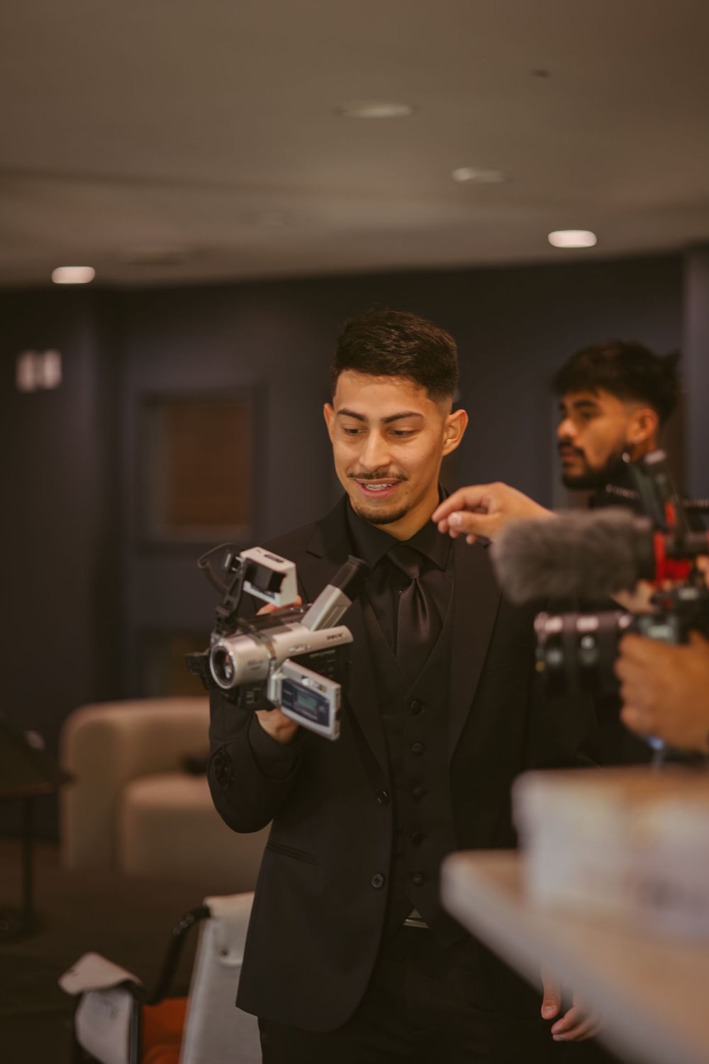 A smiling young man in a black suit holds a camera while being filmed at an indoor event.
