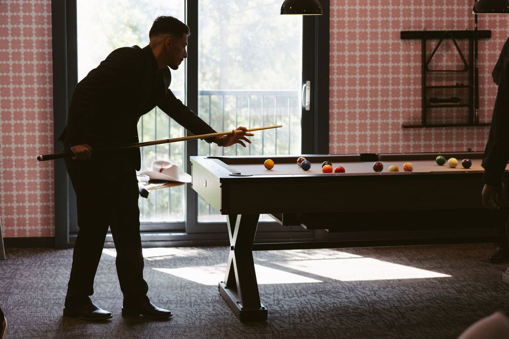 Man in black suit playing pool, leaning over billiard table with cue stick in a stylish game room.