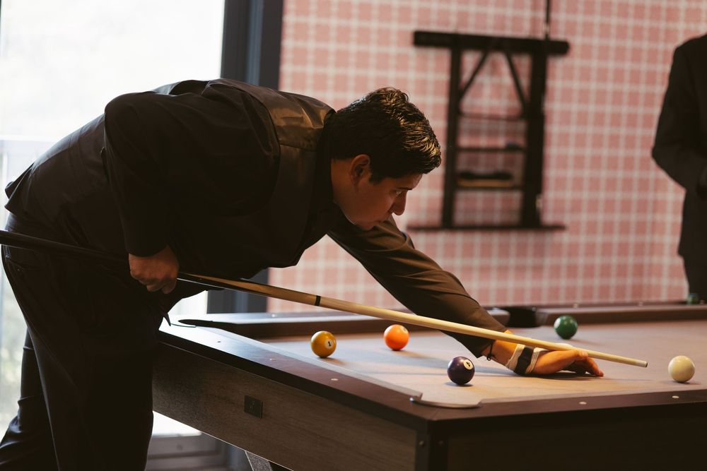 Man in black suit leaning over a pool table, carefully aiming a cue stick at colorful billiard balls.