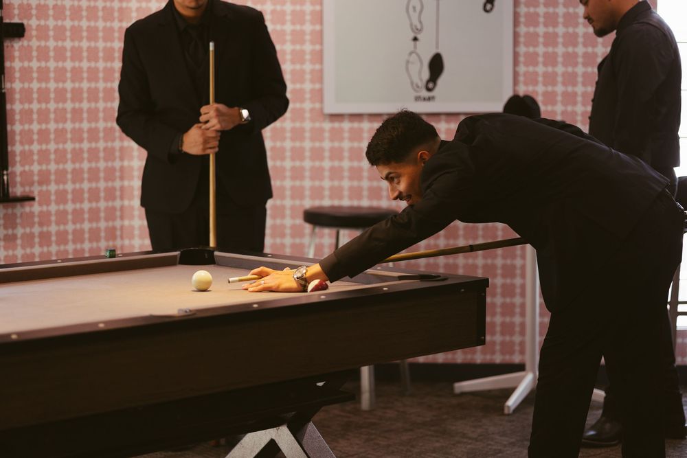 Men in black suits playing pool billiards, one leaning over table to take a shot in a pink room.
