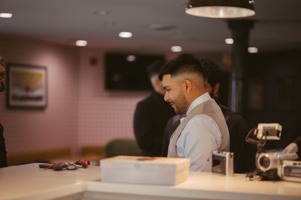 A man in a vest smiles while standing at a counter in a softly lit barbershop.