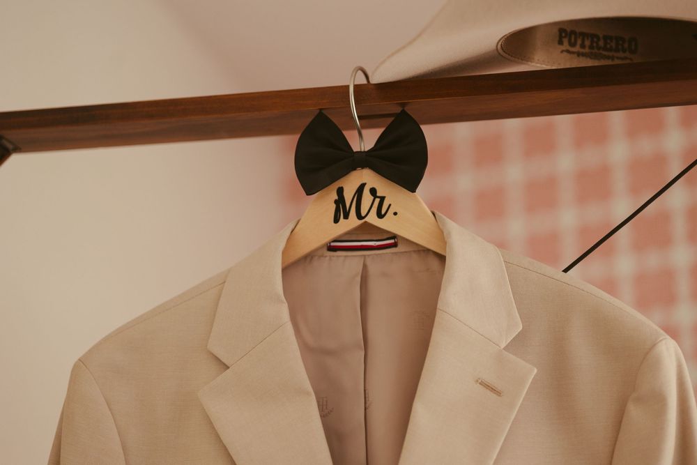 A white groom suit jacket on a wooden Mr. hanger with a black bow tie displayed on a rack.