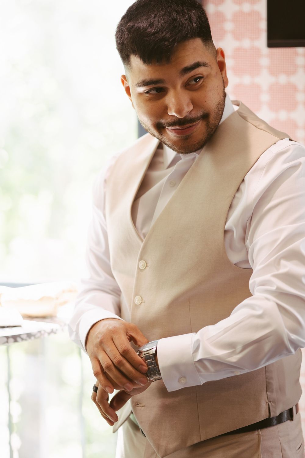 Smiling groom in white shirt and beige vest adjusting his watch, preparing for wedding ceremony indoors.