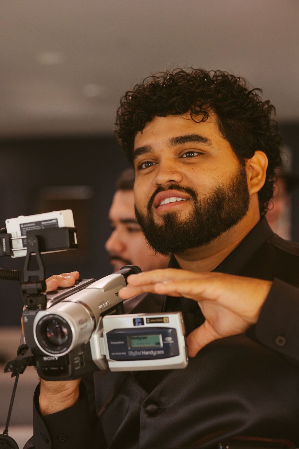 A young bearded man with curly hair smiles while holding a Sony Digital Handycam video camera at an event.