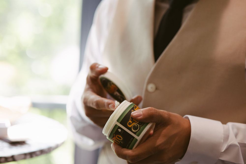 Groom in white dress shirt and vest opening a small decorative tin container during wedding preparations.