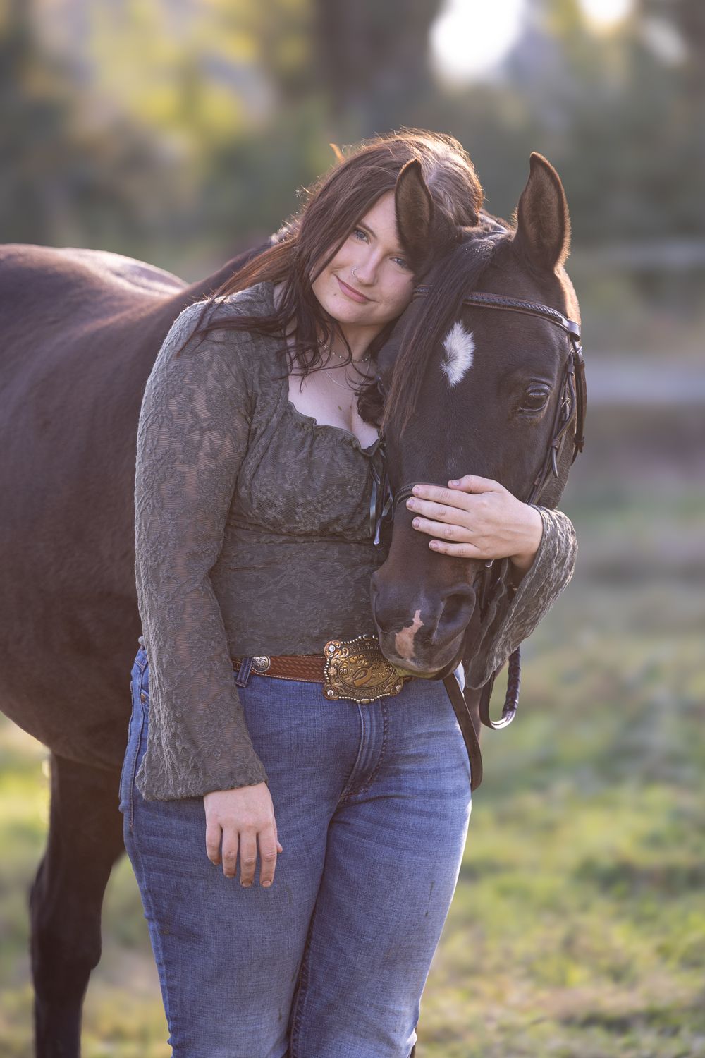 A woman with dark hair in jeans and gray top affectionately embraces a dark brown horse with a white mark outdoors.