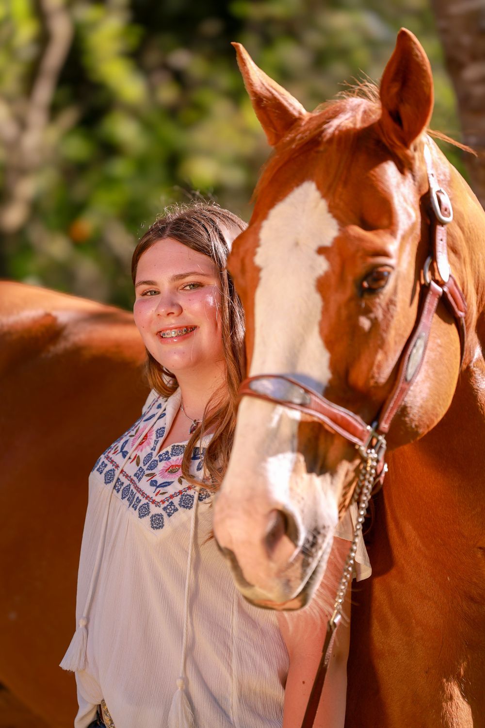 A smiling young girl poses beside a chestnut horse with a white blaze, wearing a floral top outdoors.