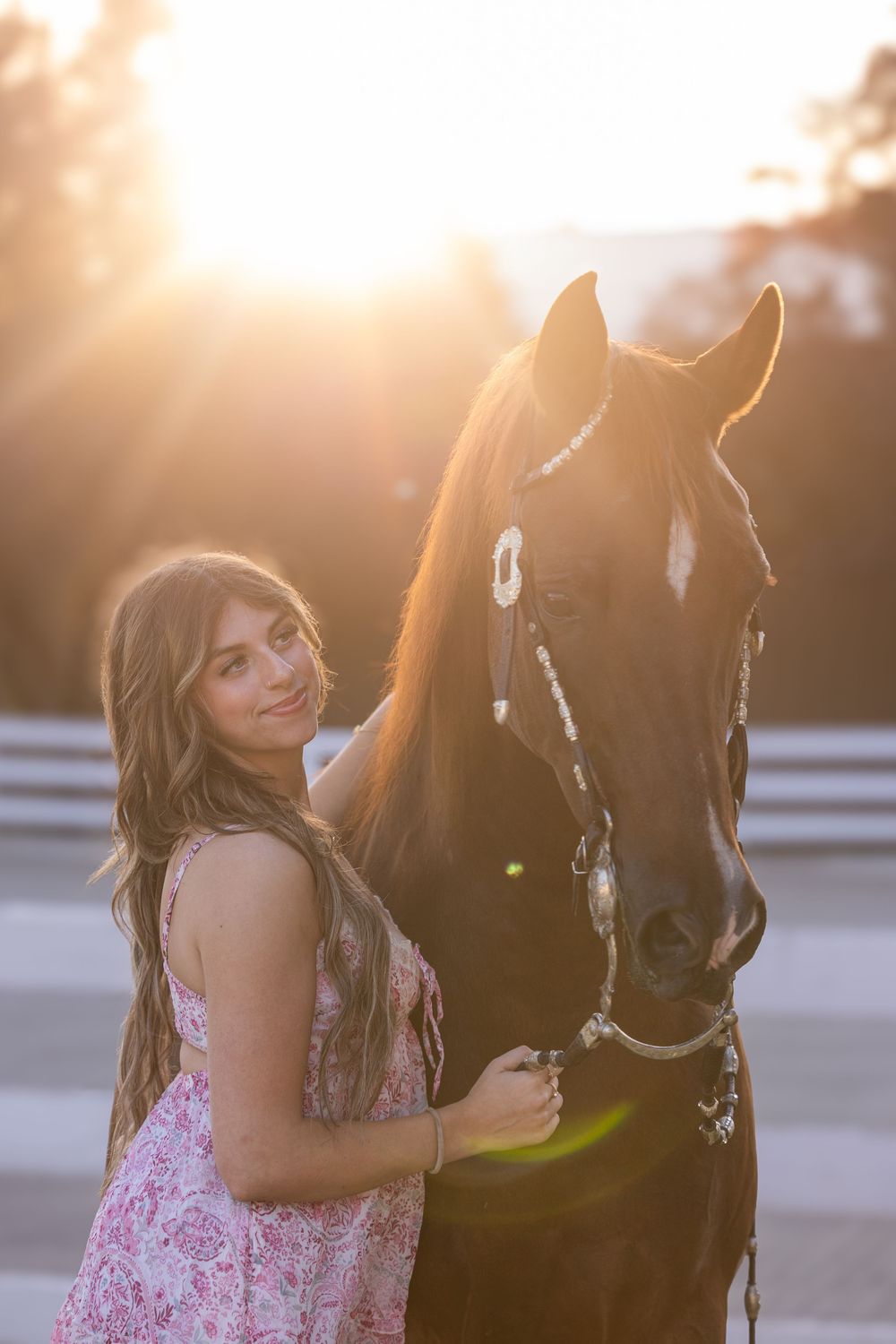 Young woman in floral dress poses with dark horse at golden sunset, warm backlight glowing behind them.