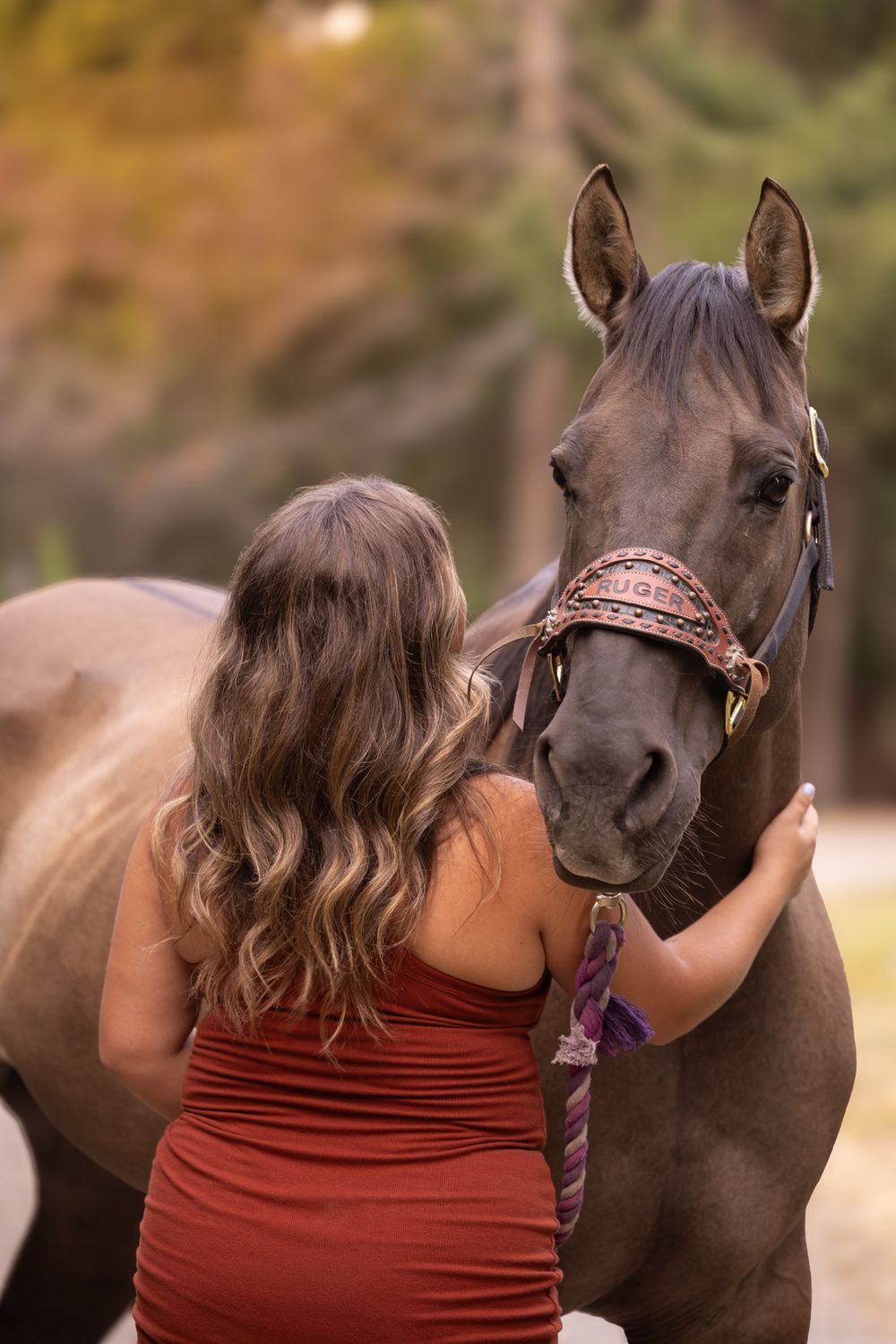 Woman in red dress tenderly embracing a dark brown horse with decorative halter in an autumn forest setting.