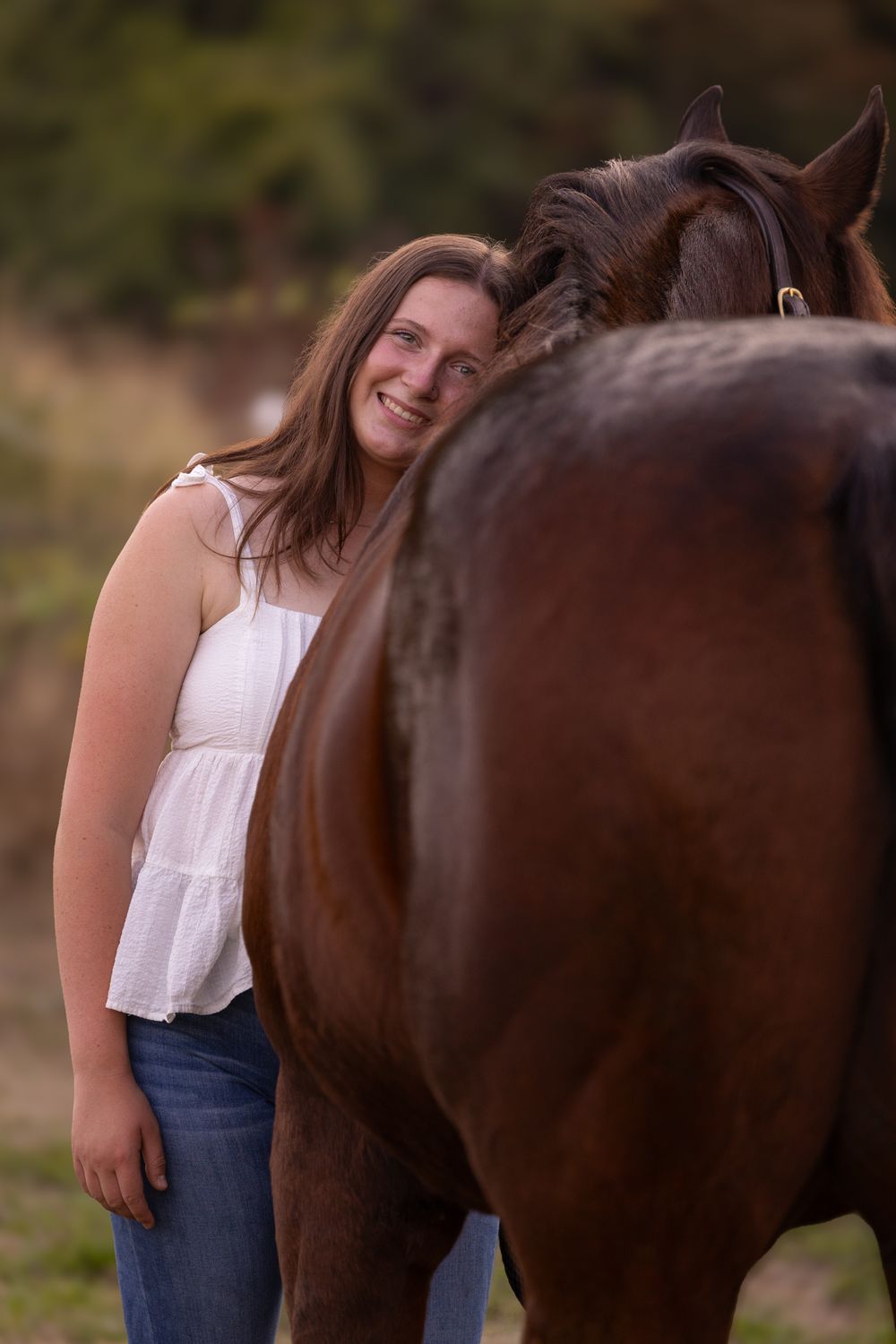A smiling young woman in a white top and jeans leans affectionately against a brown horse outdoors.