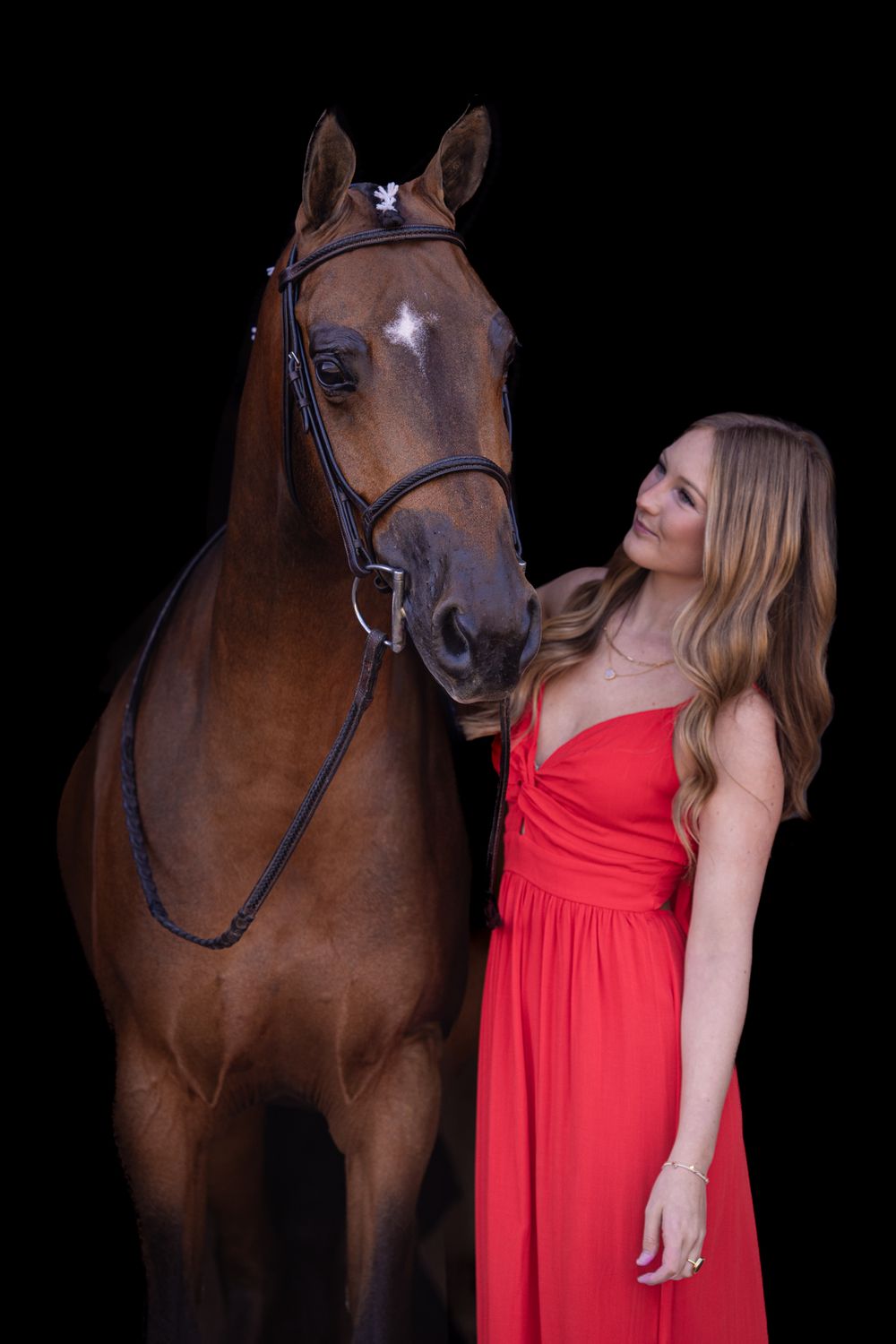 A young woman in a red gown poses with a bay horse against a black background.