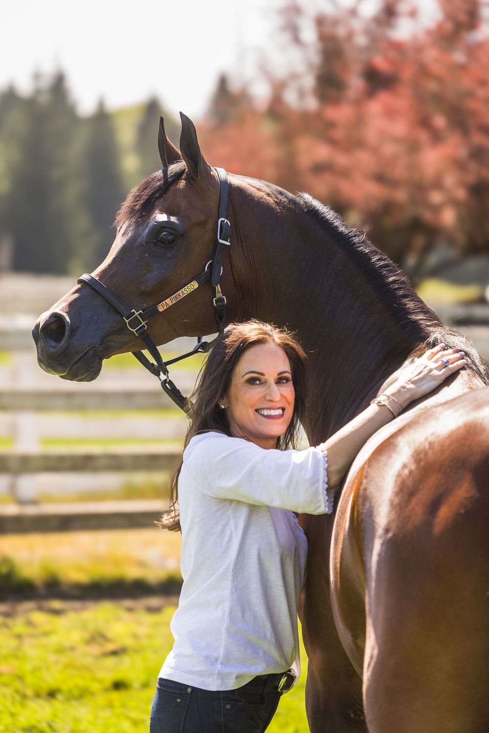 A smiling woman with long brown hair embraces a dark brown horse outdoors at a sunny equestrian farm.