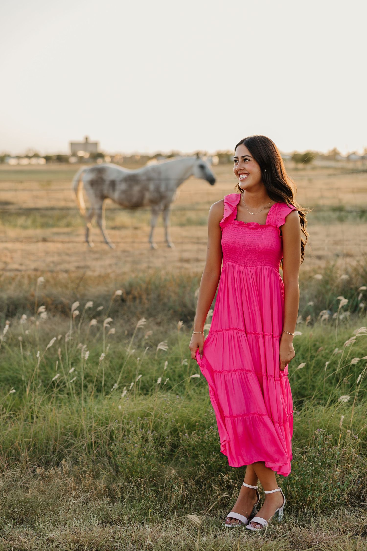 Senior Portraits in Tulia Texas wearing pink dress and posing in front of horses in a green open field.