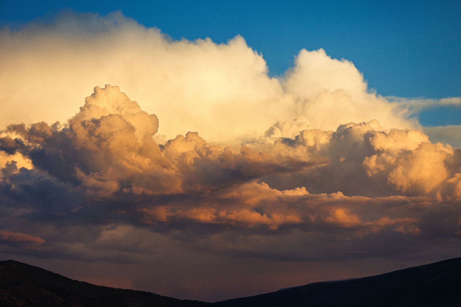 Dramatic sunset clouds with golden and orange hues over a dark silhouetted hillside landscape.