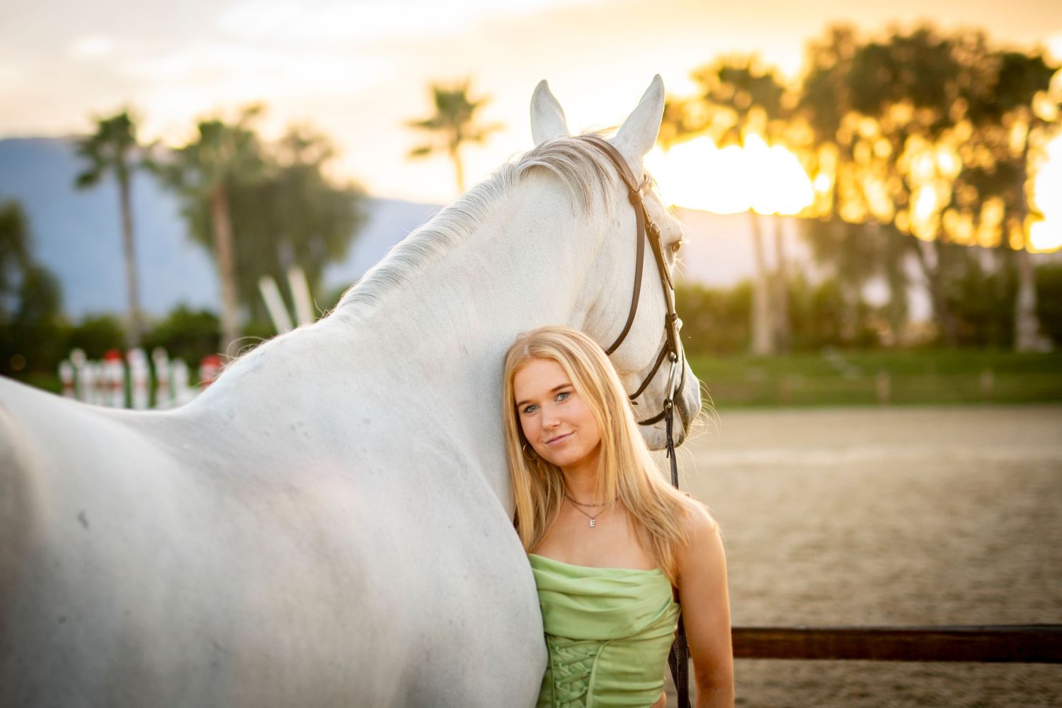 Young blonde woman posing with a white horse at sunset in an outdoor equestrian setting.