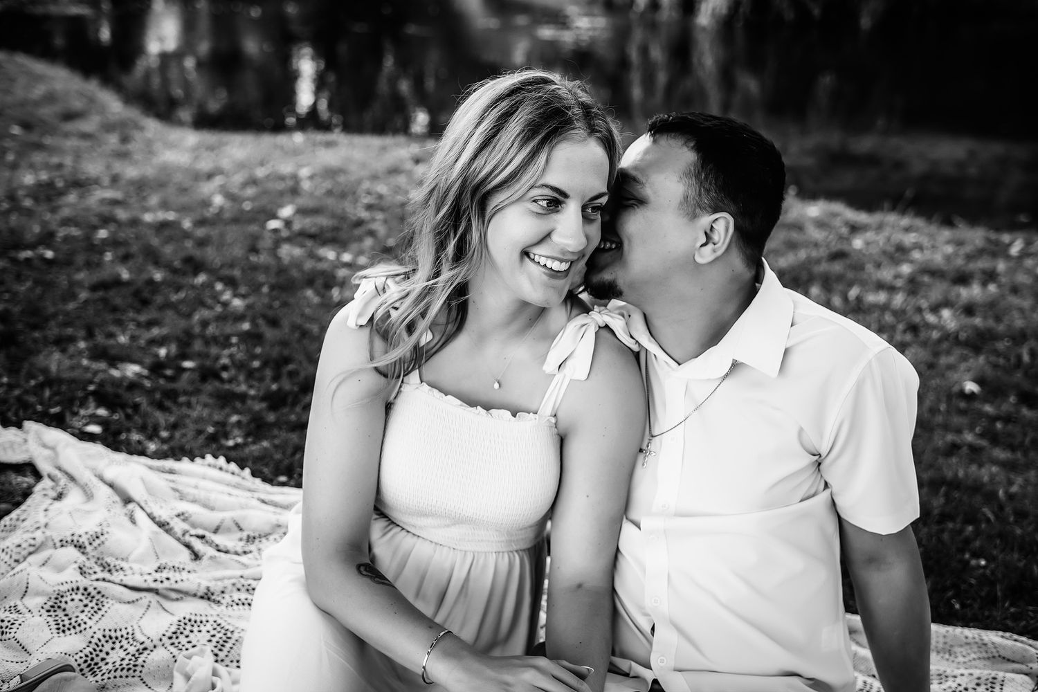 Black and white photo of a smiling couple sitting on a blanket outdoors, man whispering in womans ear.