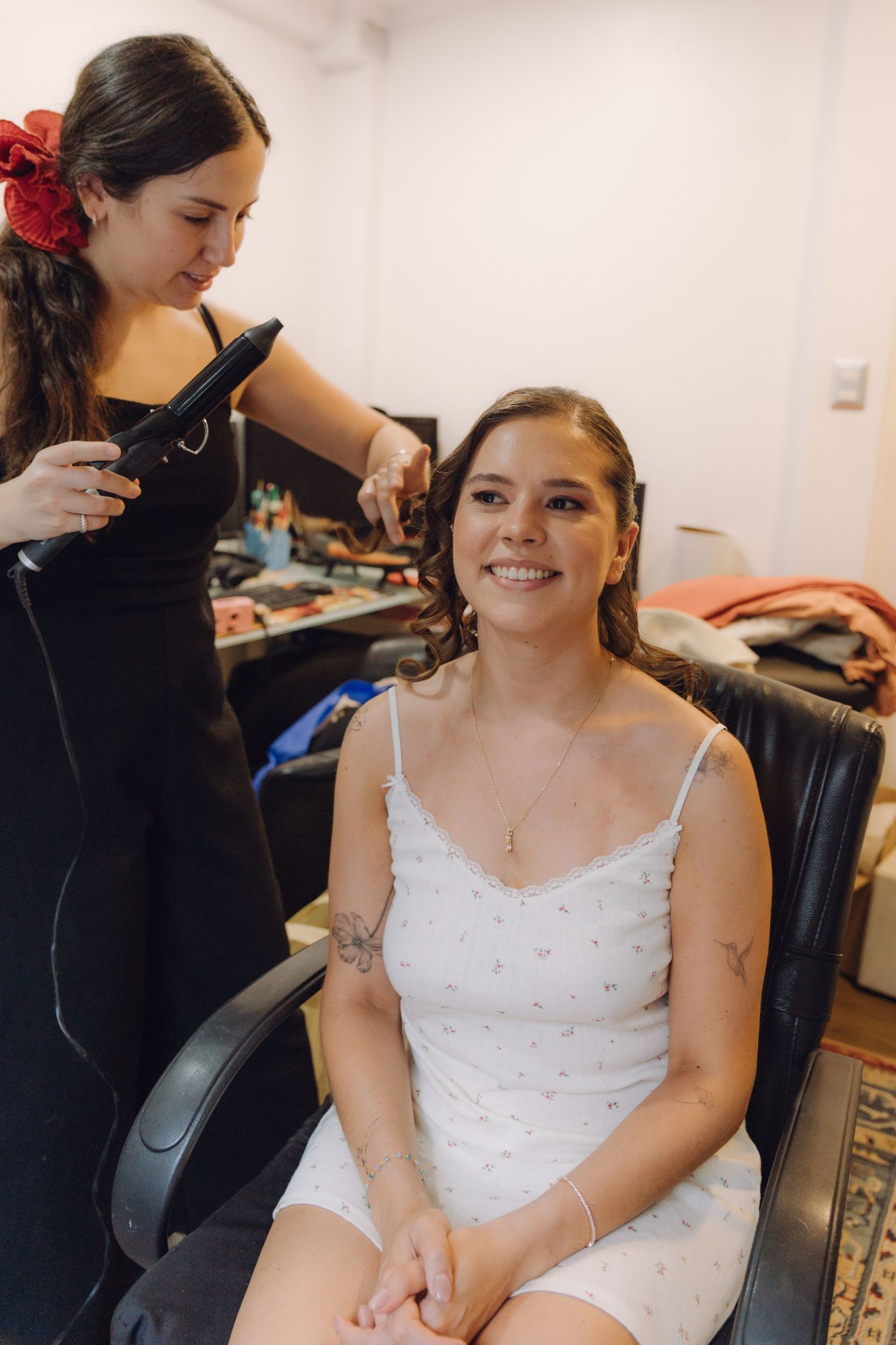 A smiling bride in a white dress gets her hair styled by a hairdresser before her wedding.