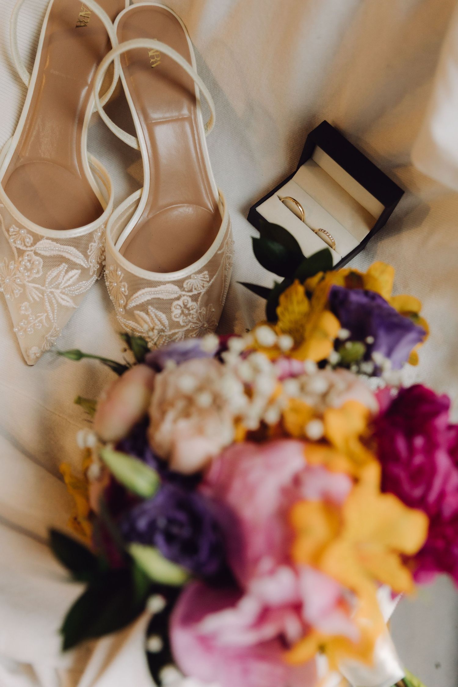 Bridal flat shoes with floral embroidery beside a ring box and colorful wedding bouquet on white fabric.
