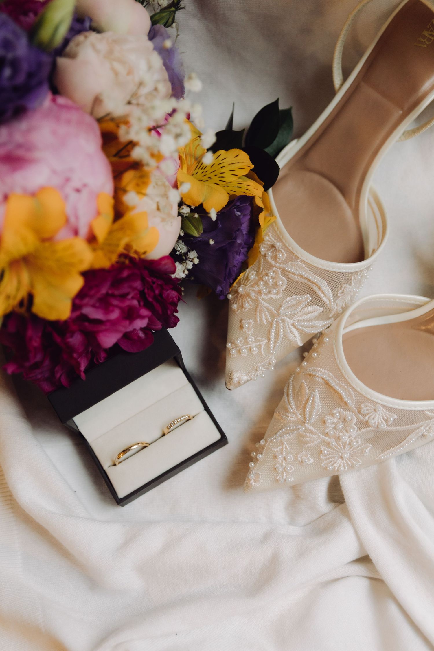 Wedding rings in black box beside lace bridal shoes and vibrant colorful bouquet on white fabric.