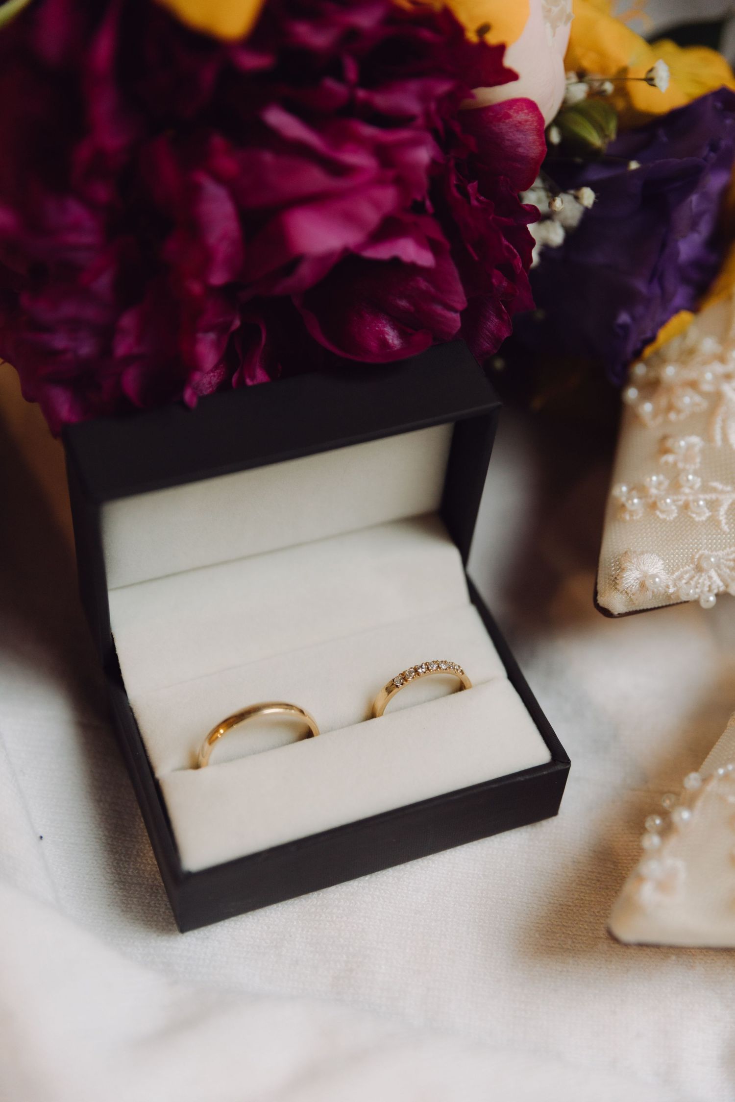 Two gold wedding bands displayed in a black ring box beside purple flowers and lace bridal shoes.