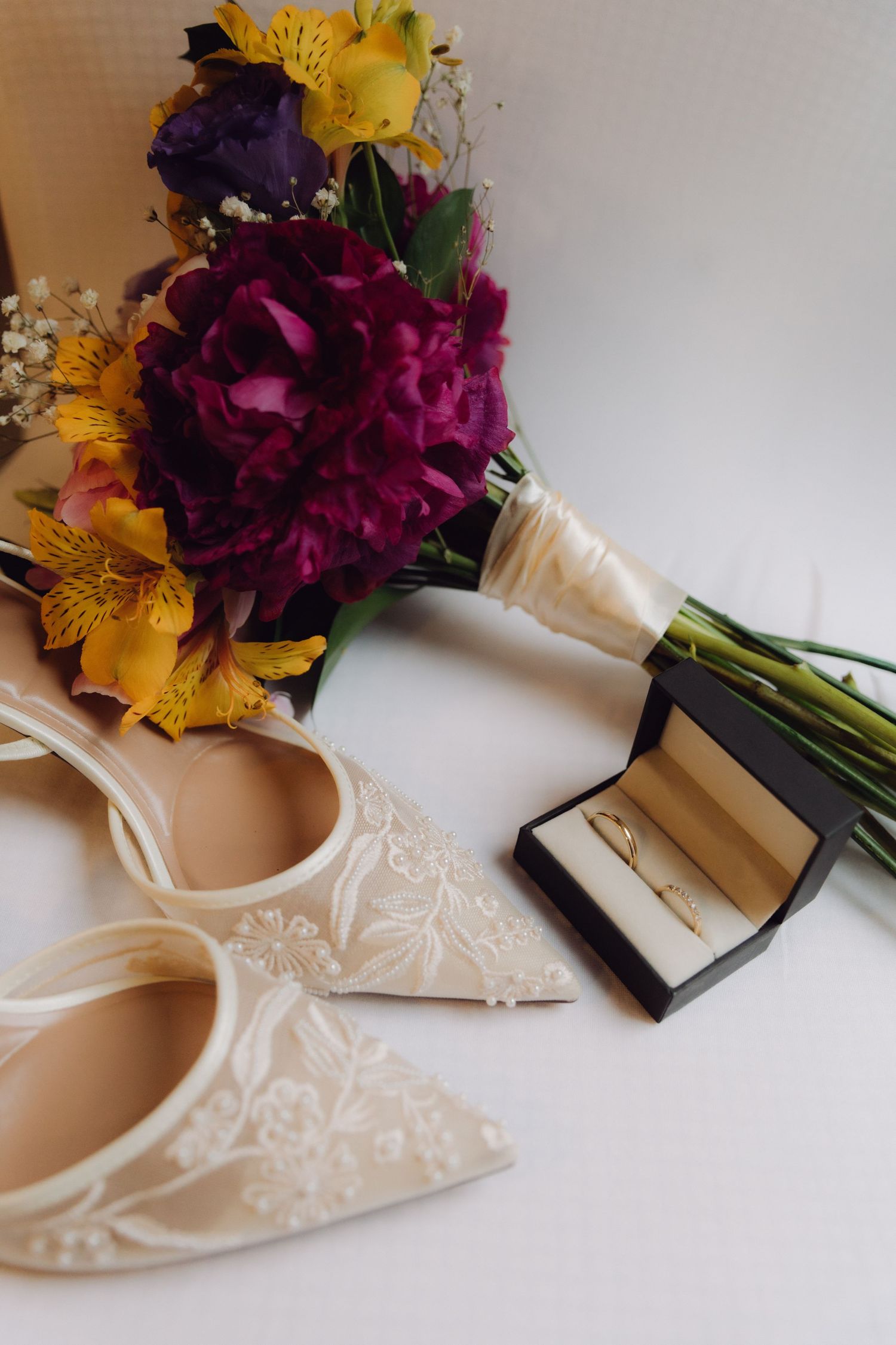 Lace bridal flats beside colorful wedding bouquet with purple and yellow flowers and gold ring box.