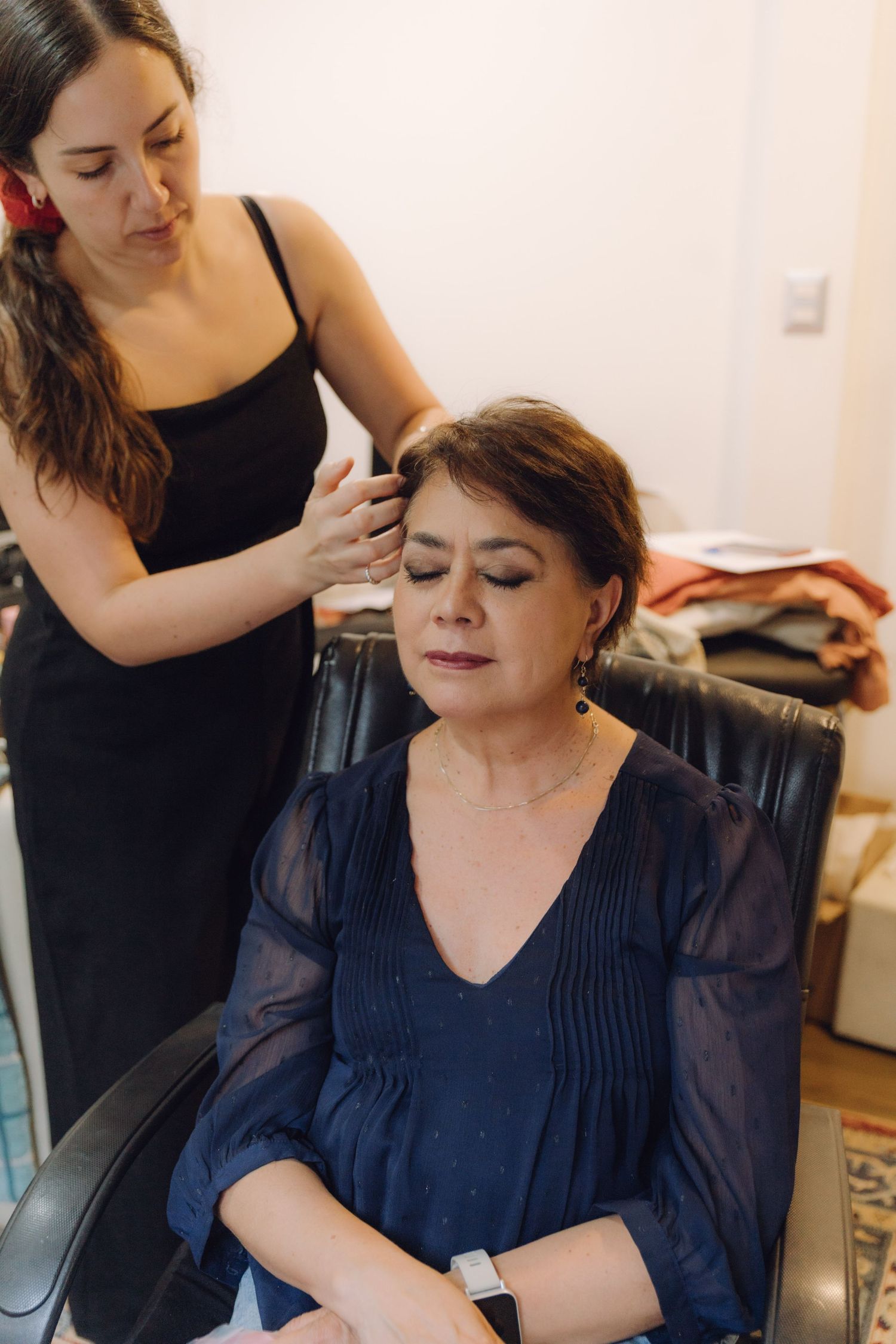 Hair stylist in black dress styling older woman with brown hair in navy blue blouse for wedding.