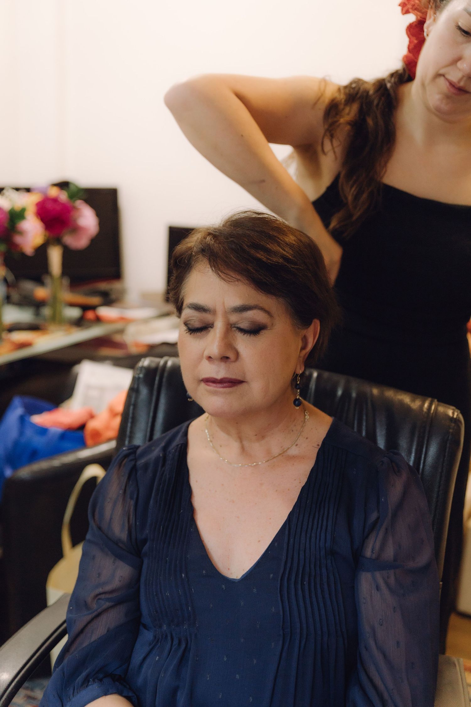 Professional hair stylist finishing elegant updo on mature woman in navy blue dress for wedding day.