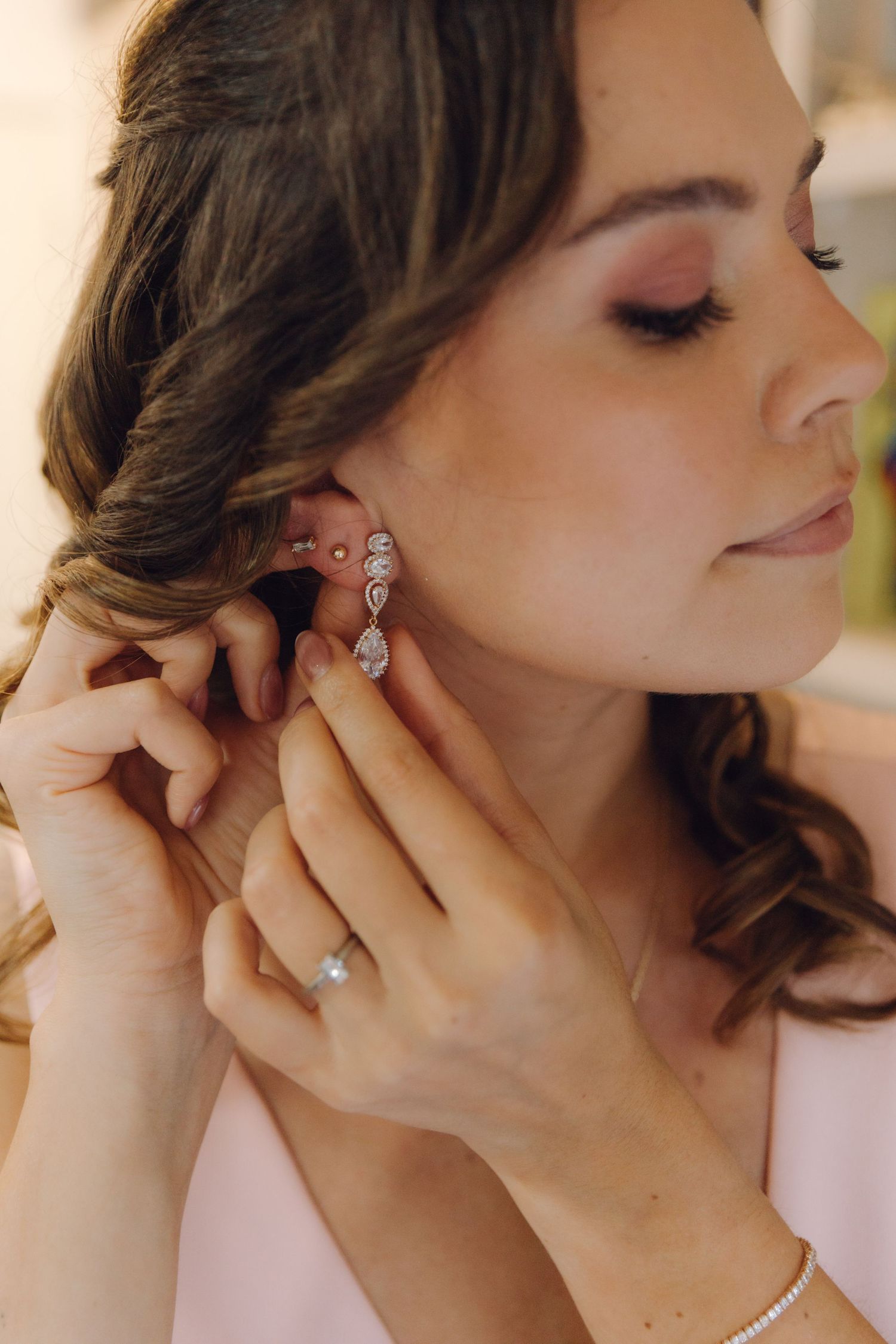 Bride fastening elegant crystal drop earring while wearing pink robe and diamond engagement ring.
