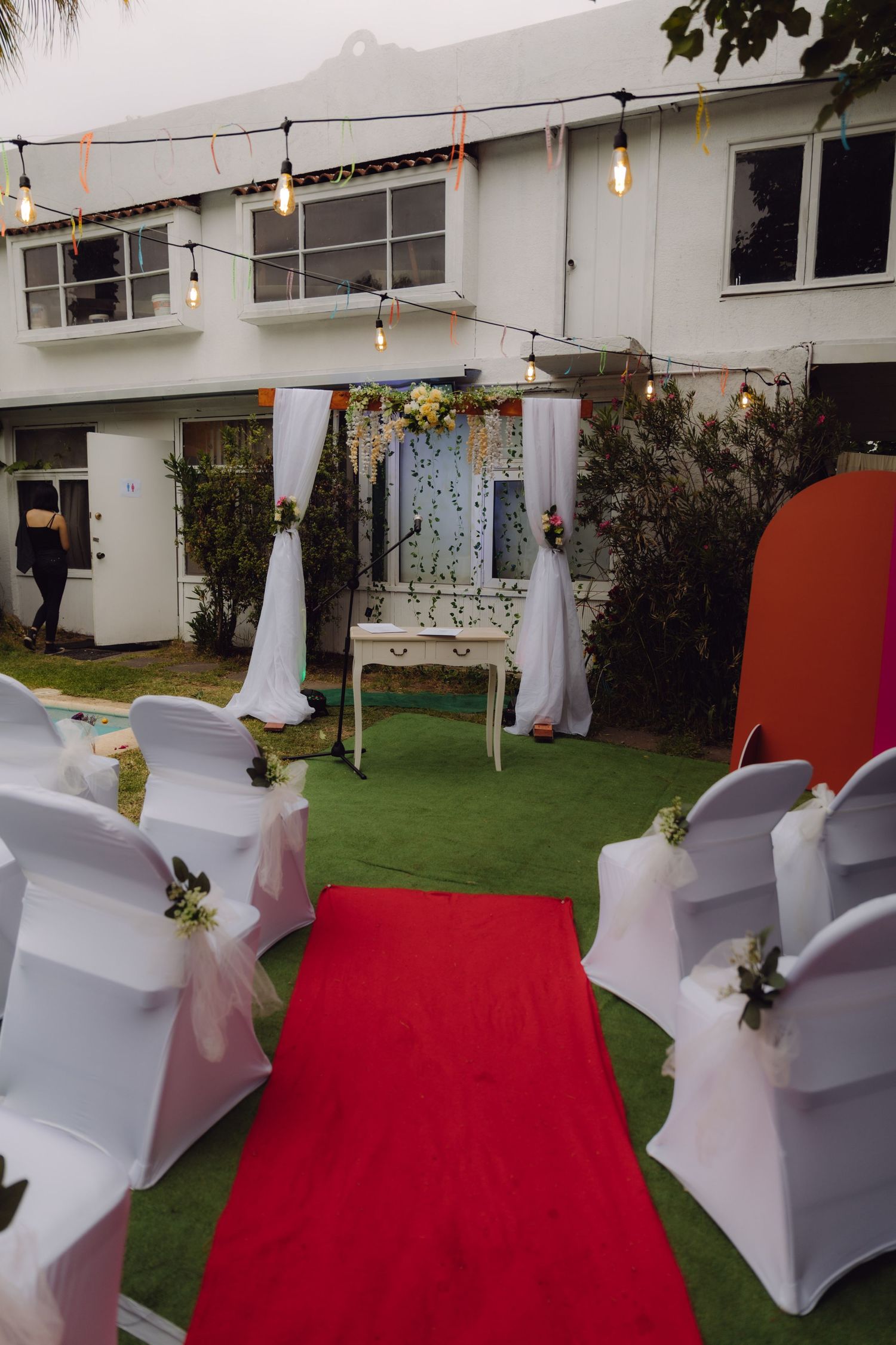 Outdoor wedding ceremony setup with red carpet aisle, white chair covers, floral arch and string lights.